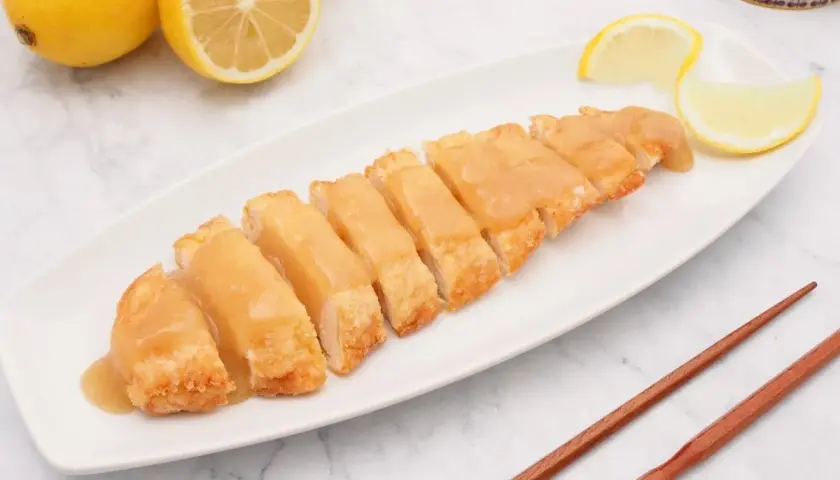 Pollo al limón chino servido en una bandeja blanca, con limones frescos y palillos de madera.