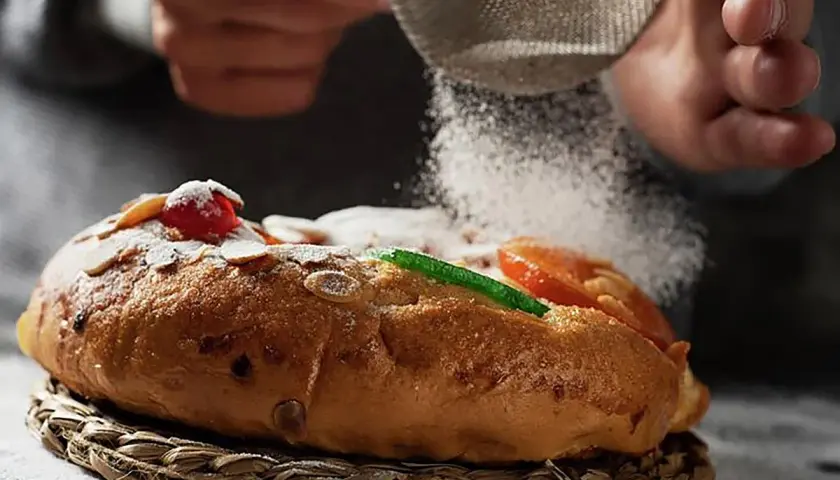 Manos espolvorean azúcar glas sobre un roscón de reyes decorado con frutas escarchadas y almendras. ¡Una delicia para tu roscón de reyes receta!