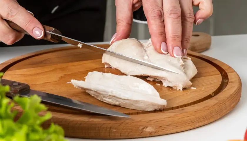 Manos cortando pechuga de pollo hervido en tabla de madera. El tiempo de cocción del pollo hervido es clave para su jugosidad.