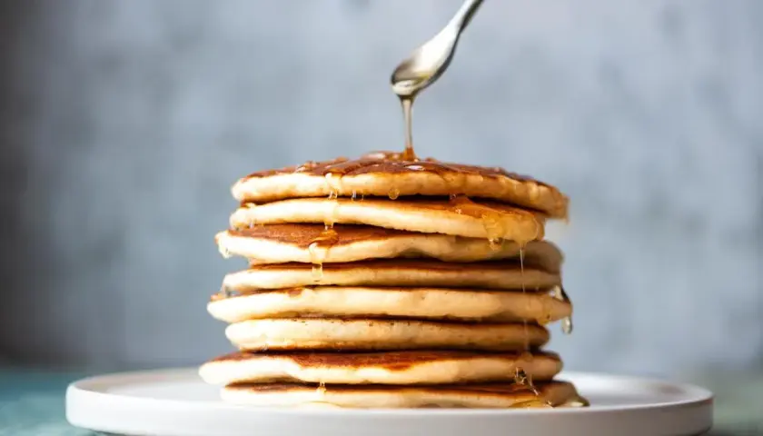 Una pila de tortitas sin mantequilla, doradas y esponjosas, con sirope cayendo de una cuchara.