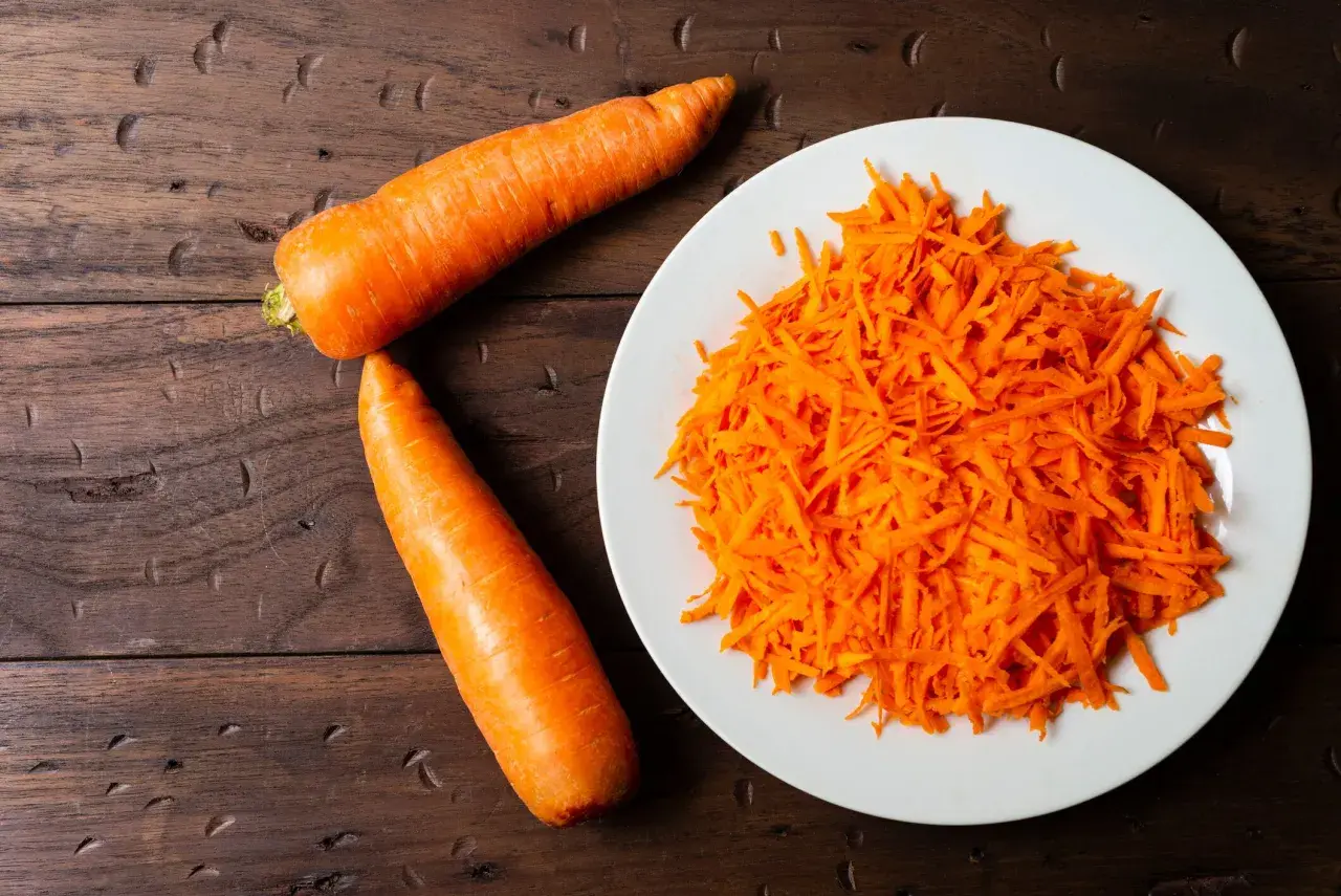 Dos zanahorias enteras y una porción de zanahoria rallada en un plato blanco. Preparando ingredientes para conocer el tiempo coccion zanahoria.