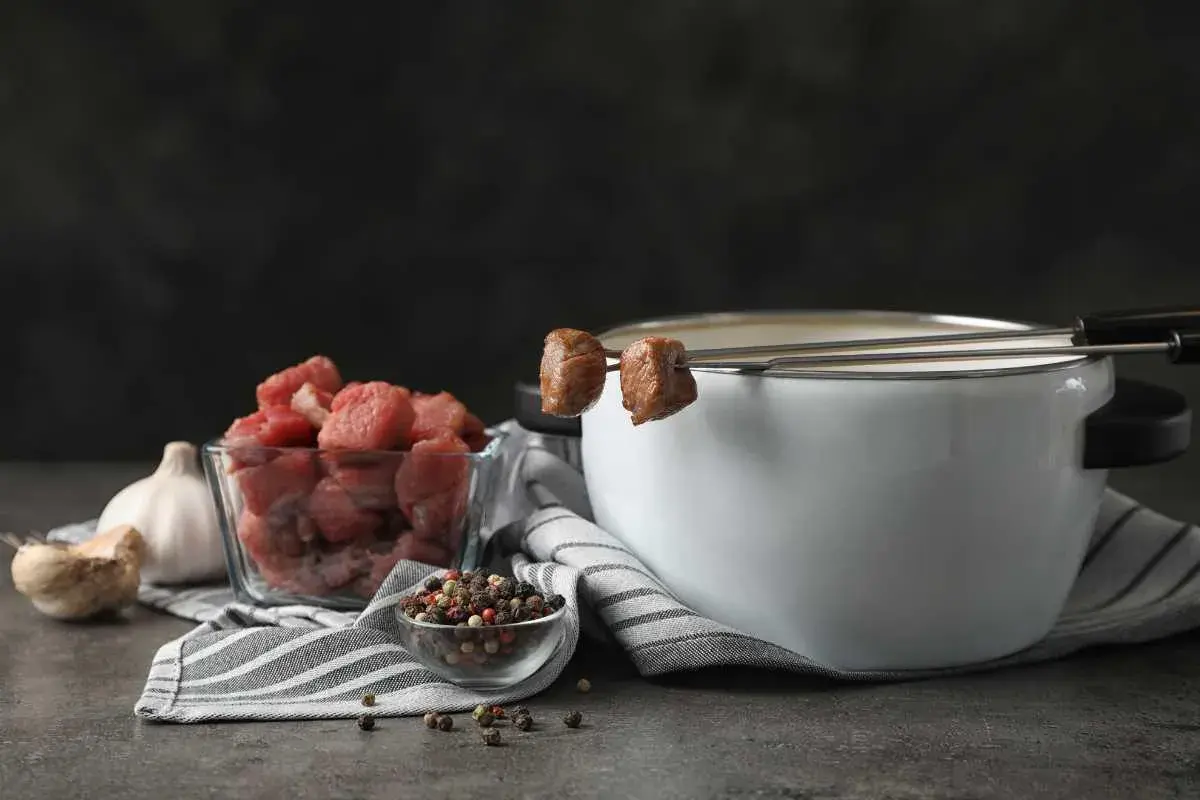 Preparación para un delicioso fondue de carne: trozos de carne cruda, ajo, granos de pimienta y una olla con caldo humeante.