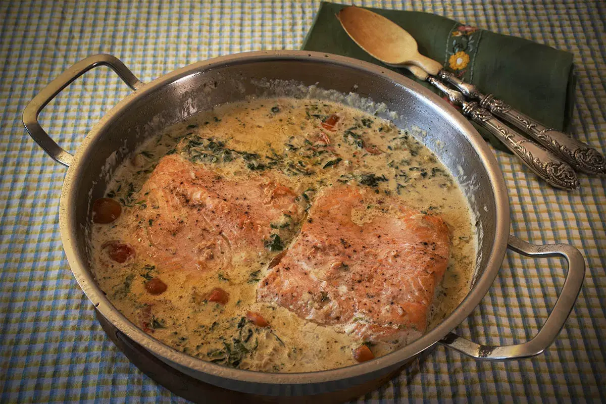 Dos filetes de salmón a la toscana en una sartén con salsa cremosa, espinacas y tomates cherry.