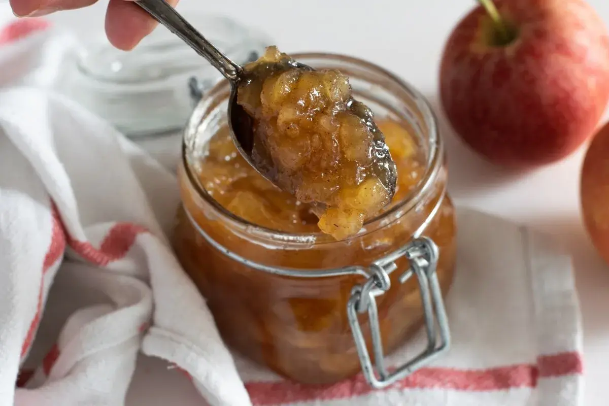 Cuchara sirviendo mermelada de manzana casera de un frasco. Manzanas rojas frescas al lado.