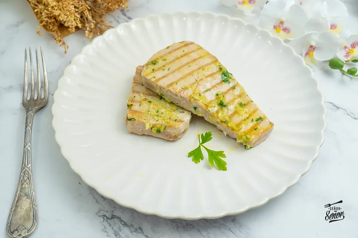Dos filetes de atún a la plancha, con marcas de parrilla y aderezados con ajo y perejil, servidos en un plato blanco con un tenedor.