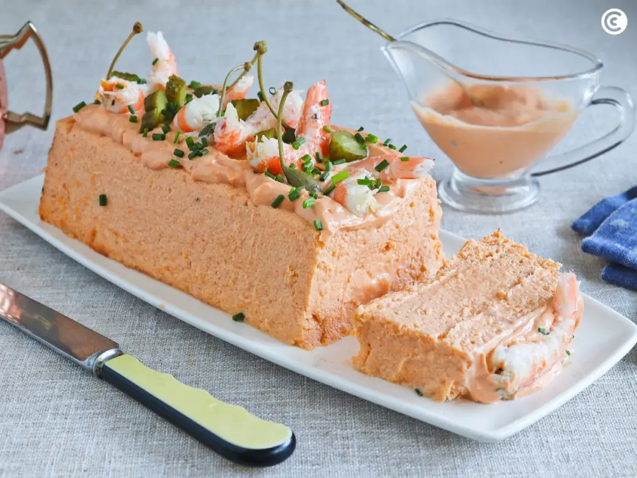 Pastel de pescado decorado con gambas, pepinillos y alcaparras. Servido en una bandeja blanca con una porción cortada.