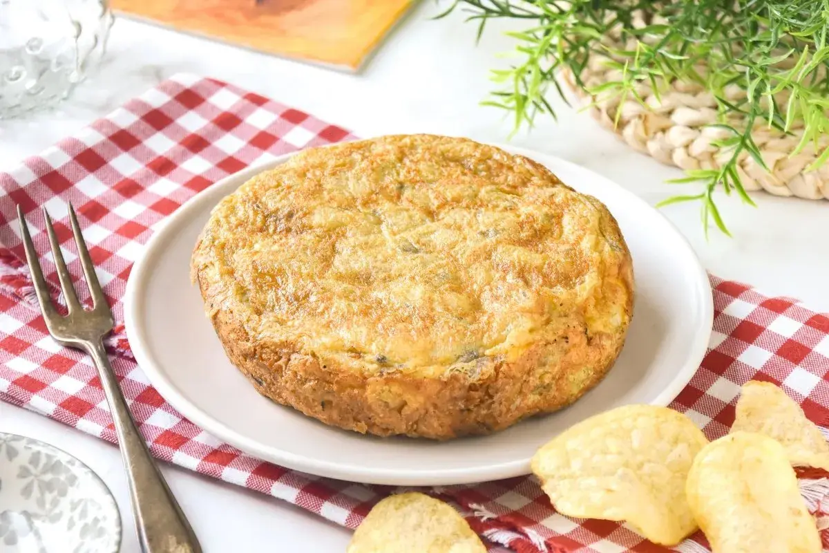 Tortilla con patatas de bolsa, dorada y jugosa, servida en plato blanco sobre mantel de cuadros rojos. Patatas fritas crujientes alrededor.