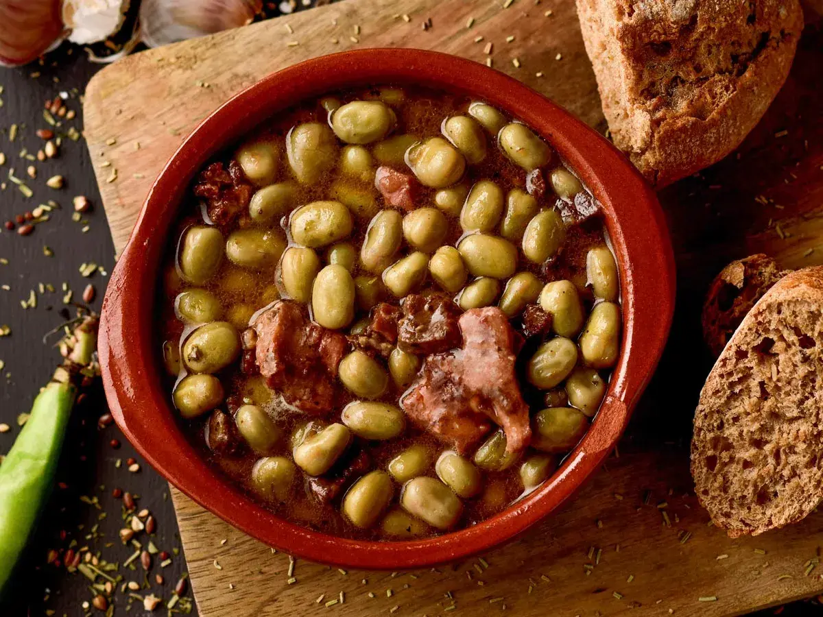 Un cuenco de barro con habas a la catalana, trozos de tocino y pan rústico al lado.