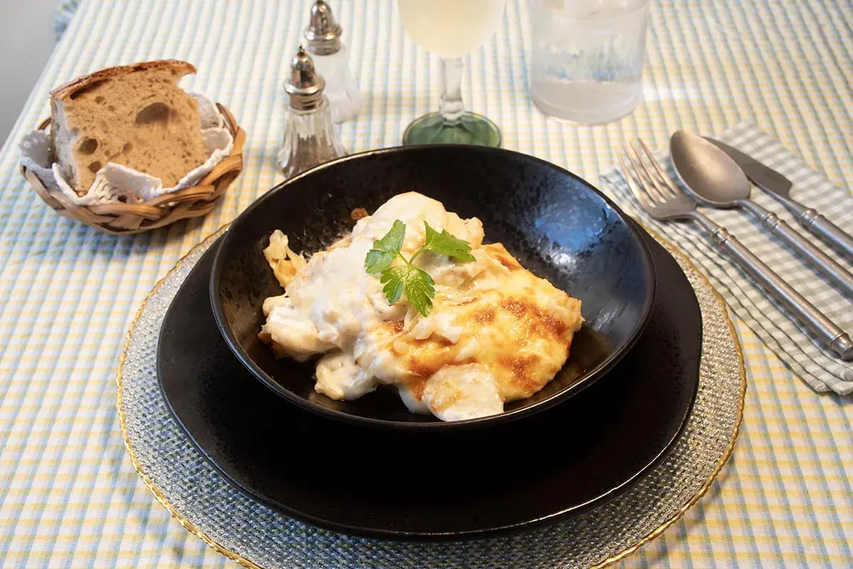 Delicioso bacalao a la nata gratinado, servido en un cuenco negro, acompañado de pan rústico y cubiertos.