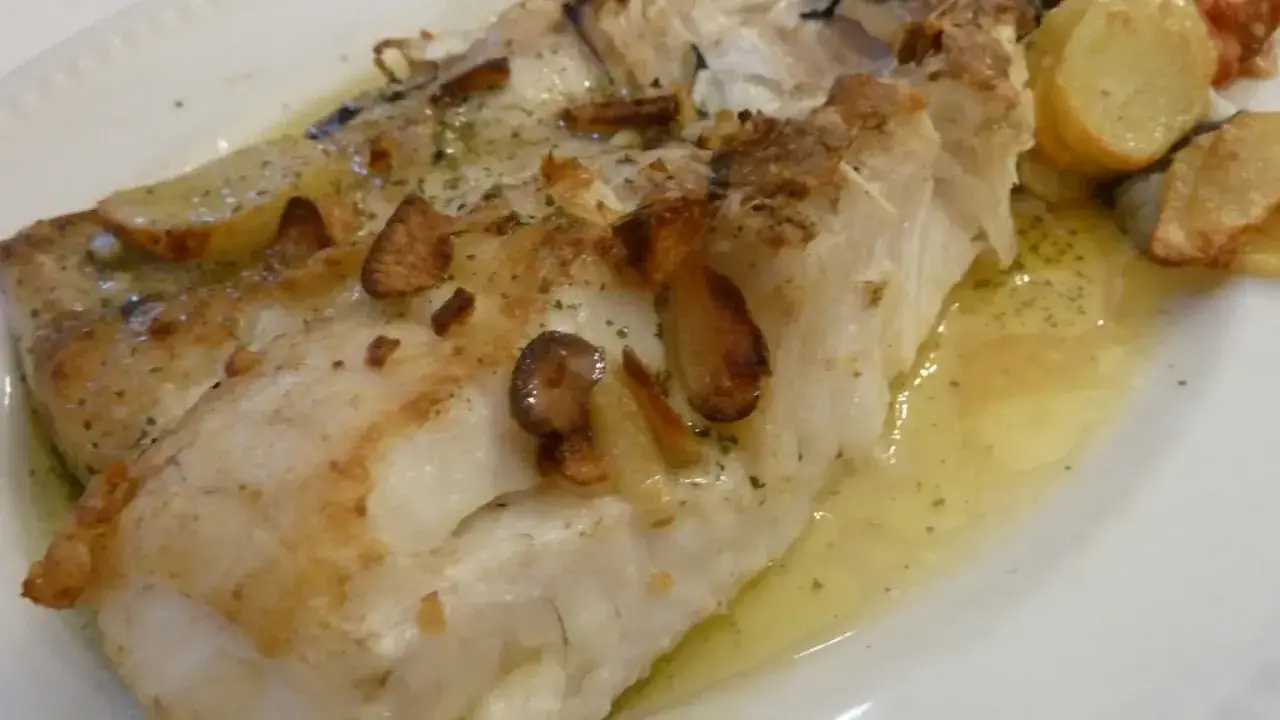 Merluza al horno sin patatas, jugosa y dorada, con láminas de ajo crujientes y hierbas aromáticas. Un plato ligero y sabroso.