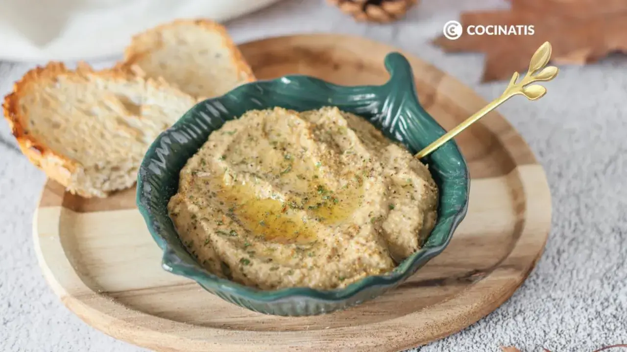 Delicioso pate de berenjena cremoso, servido en un cuenco verde con un toque de aceite de oliva y perejil. Acompañado de tostadas crujientes.