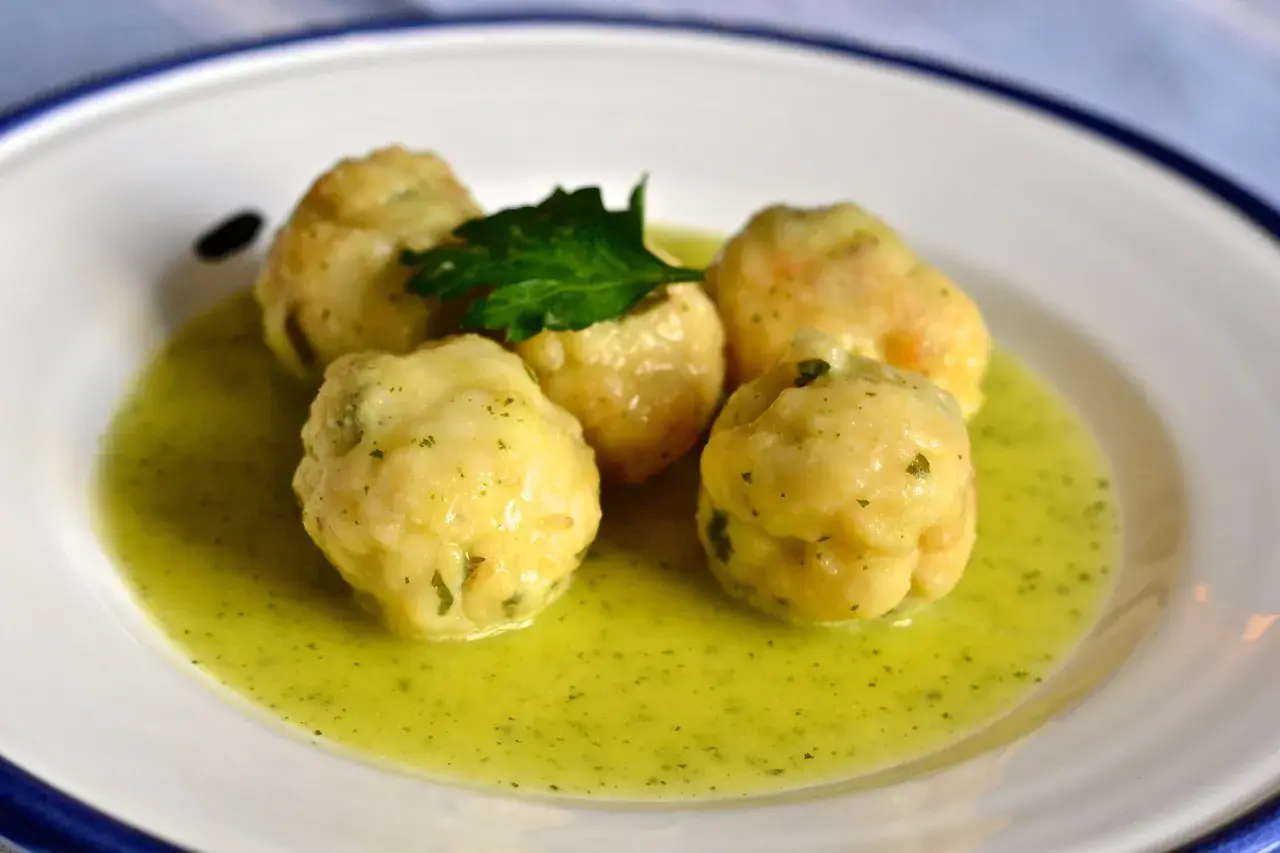 Cinco albóndigas de merluza caseras flotan en una salsa verde cremosa, adornadas con una ramita de perejil fresco.