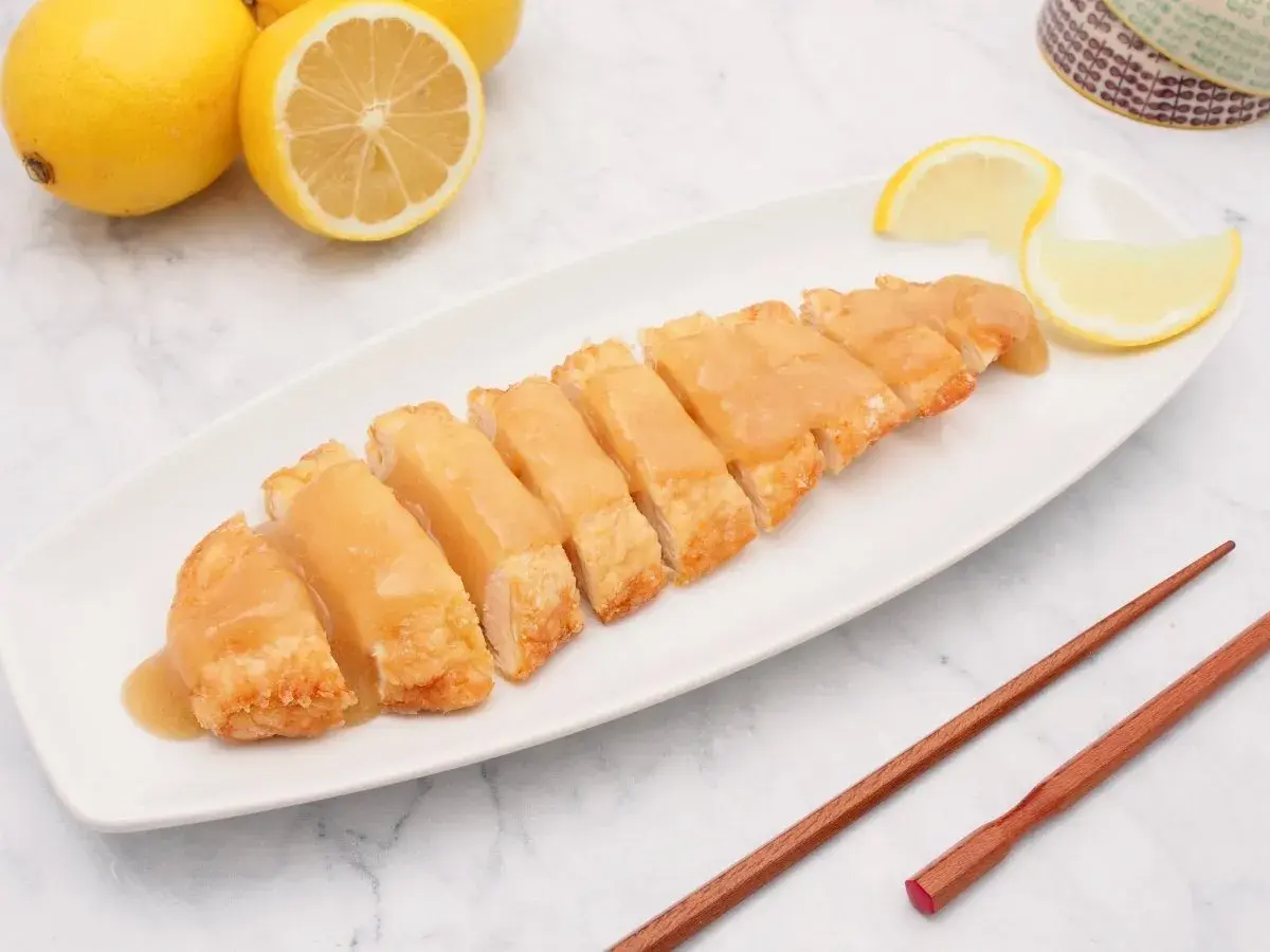 Pollo al limón chino servido en una bandeja blanca, con limones frescos y palillos de madera.