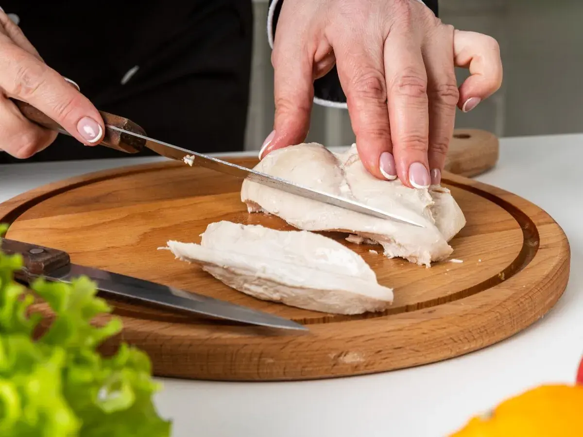 Manos cortando pechuga de pollo hervido en tabla de madera. El tiempo de cocción del pollo hervido es clave para su jugosidad.