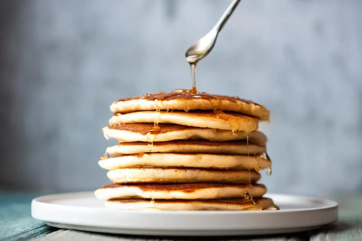 Una pila de tortitas sin mantequilla, doradas y esponjosas, con sirope cayendo de una cuchara.