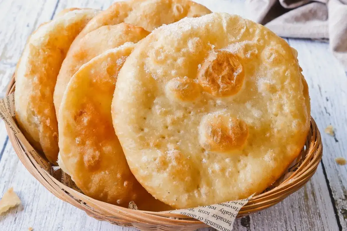 Bu&ntilde;uelos de viento dorados y esponjosos, espolvoreados con az&uacute;car, servidos en una cesta de mimbre.