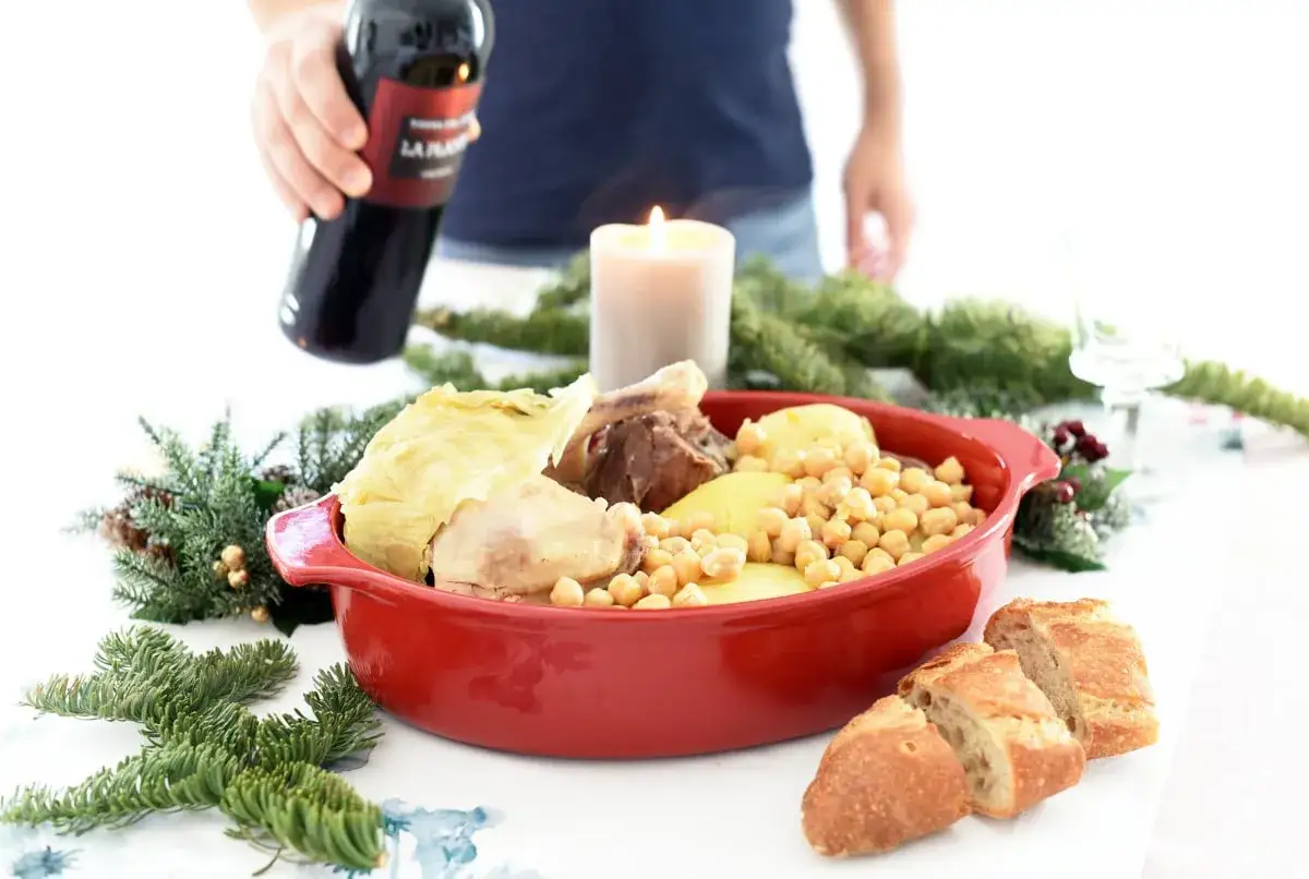 Un delicioso cocido en olla express, servido en una cazuela roja, con garbanzos, patatas, carne y repollo. Acompa&ntilde;ado de pan y vino.