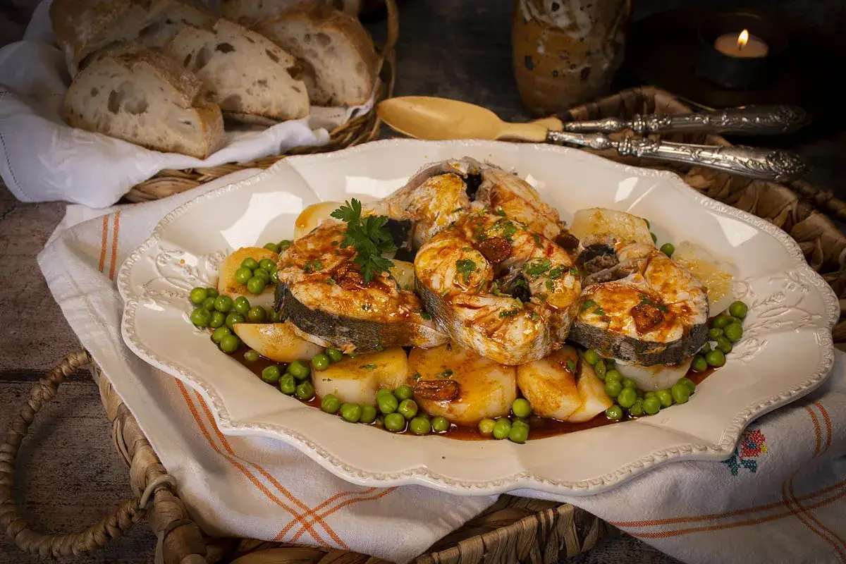 Merluza a la gallega arguiñano, jugosos trozos de pescado con patatas y guisantes, servidos en una fuente decorada.
