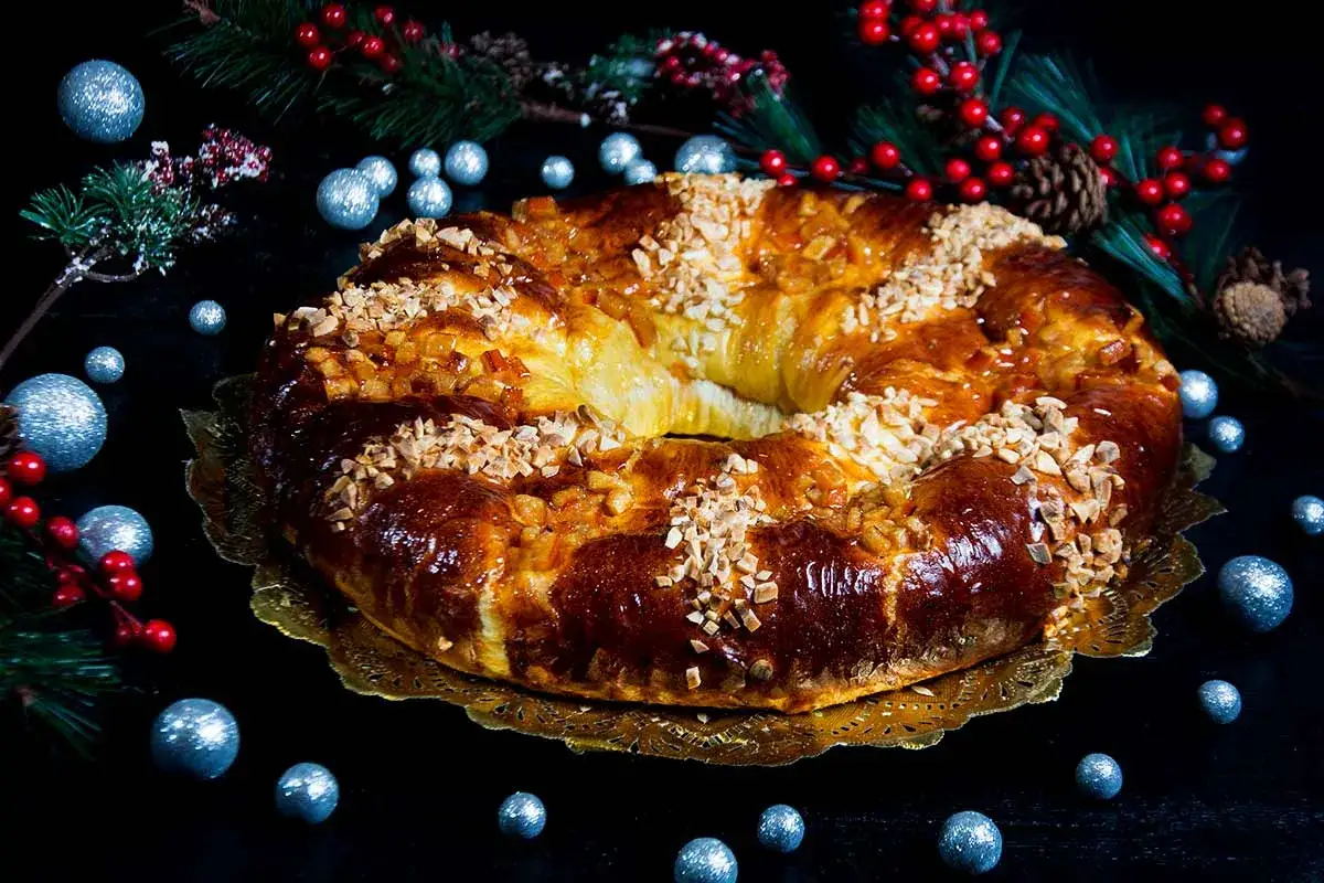Delicioso rosc&oacute;n de reyes receta, adornado con frutos secos y glaseado, rodeado de decoraci&oacute;n navide&ntilde;a.