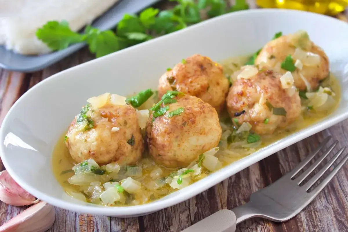 Cuatro alb&oacute;ndigas de merluza caseras en una salsa cremosa con cebolla y perejil, servidas en un plato blanco.