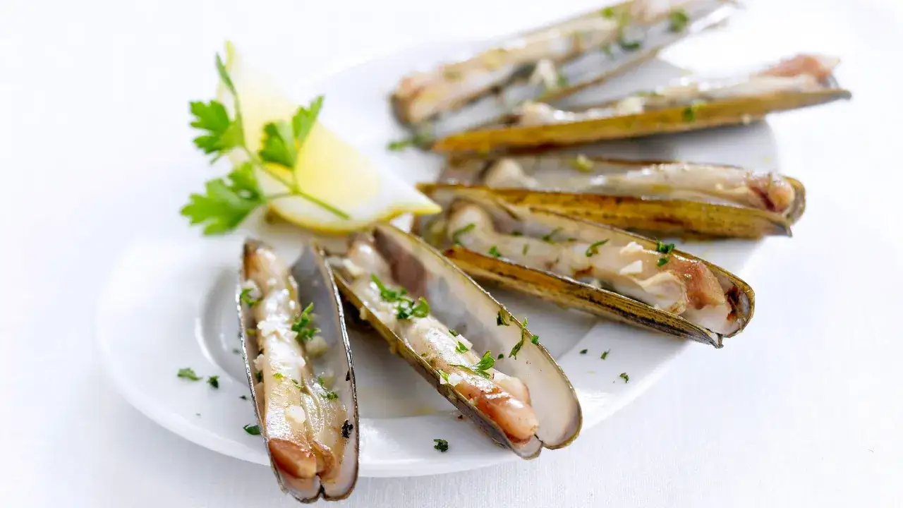 Deliciosas navajas a la plancha, servidas en una bandeja blanca con perejil picado y un gajo de limón.