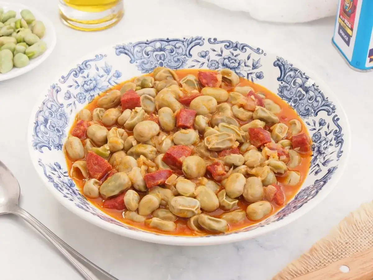 Plato de habas tiernas guisadas con trozos de chorizo espa&ntilde;ol en una salsa rojiza, servido en una vajilla azul y blanca.