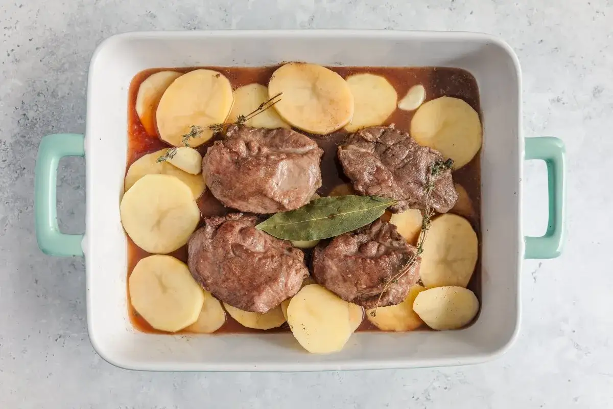 Carrilleras al horno karlos argui&ntilde;ano con patatas en rodajas y hierbas arom&aacute;ticas, listas para cocinar.