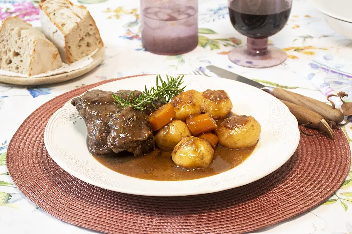 Carrilleras de ternera estofadas con patatas y zanahorias, servidas con pan rústico y vino tinto.