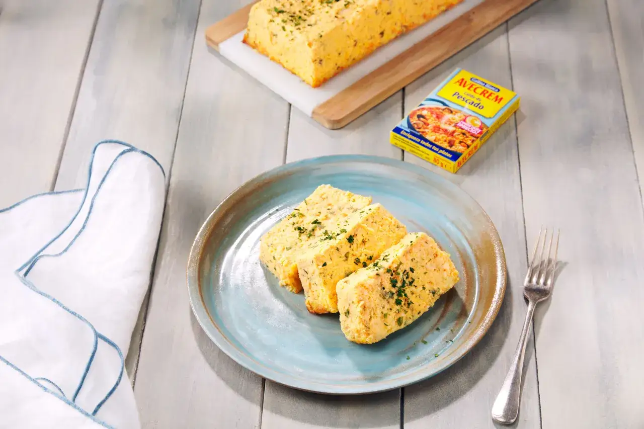 Porción de pastel de pescado, esponjoso y dorado, servido en plato azul. Al fondo, otro trozo y un paquete de caldo de pescado.