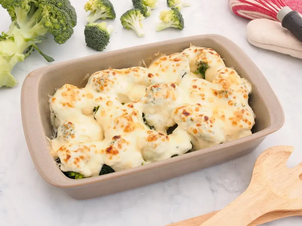 Brocoli con bechamel gratinado en un recipiente, con brócoli fresco al lado.