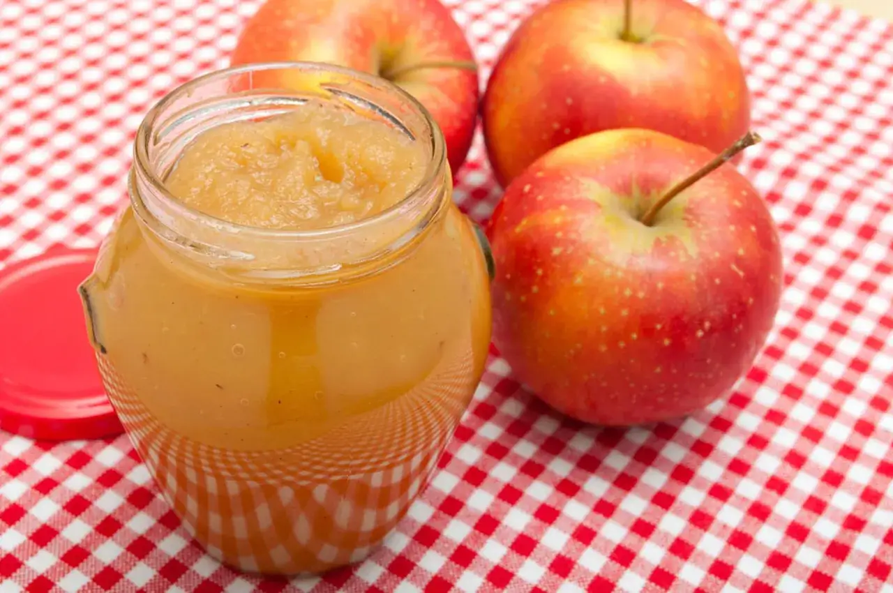 Tarro de mermelada de manzana casera junto a manzanas rojas frescas sobre un mantel a cuadros.