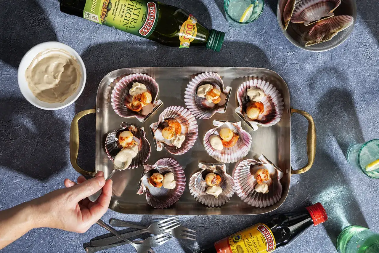 Vieiras a la plancha servidas en sus conchas, con una salsa cremosa. Una mano a&ntilde;ade m&aacute;s salsa a una de ellas.