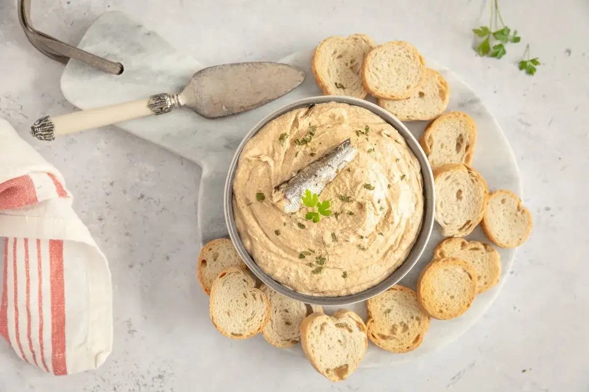 Un delicioso paté de sardinas cremoso, decorado con una sardina entera y perejil, servido con tostadas crujientes.