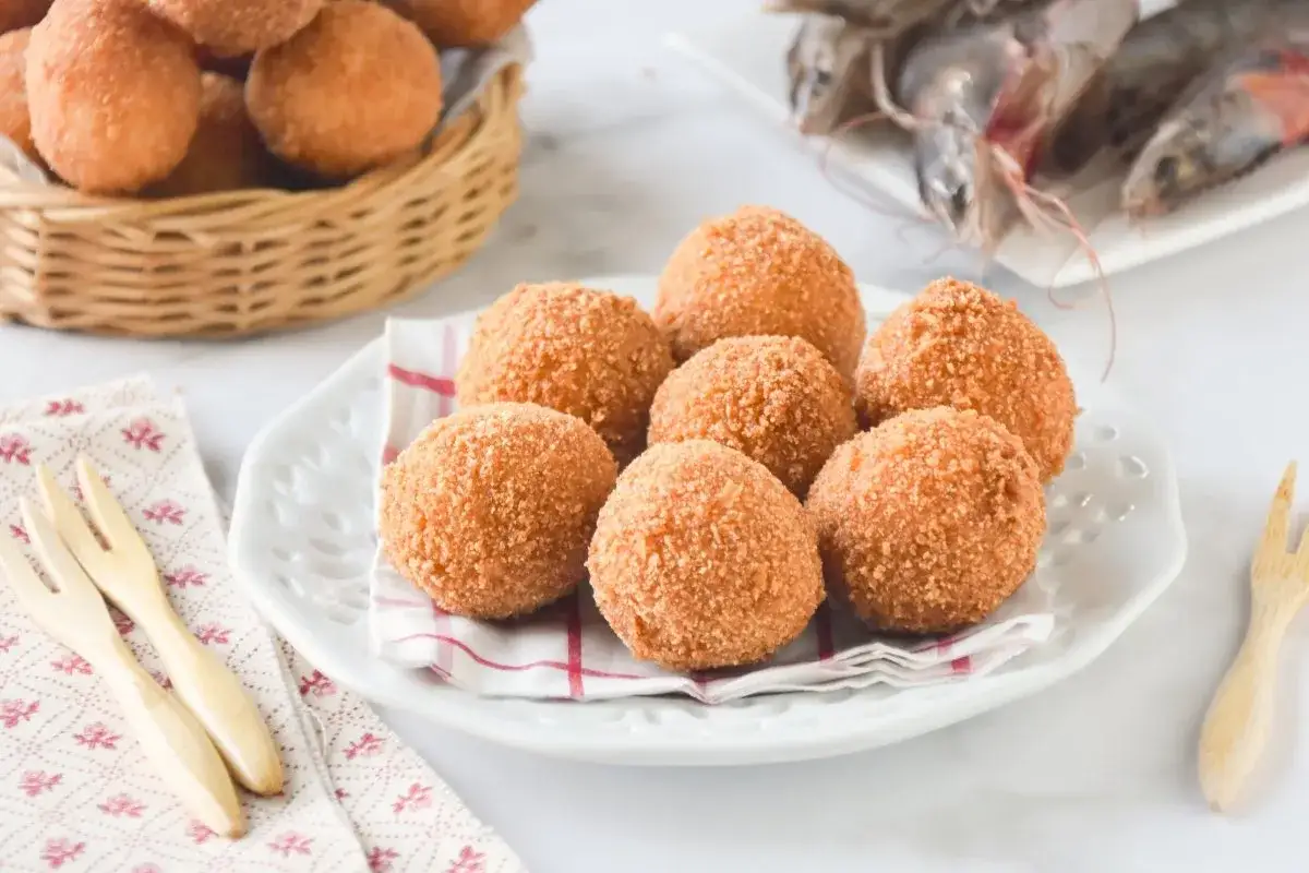 Croquetas de gambas doradas y crujientes en un plato blanco, con gambas frescas al fondo.