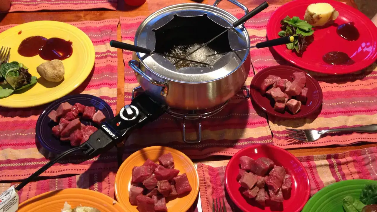 Prepara una deliciosa fondue de carne. Cubos de carne cruda esperan en platos de colores junto a una olla burbujeante.
