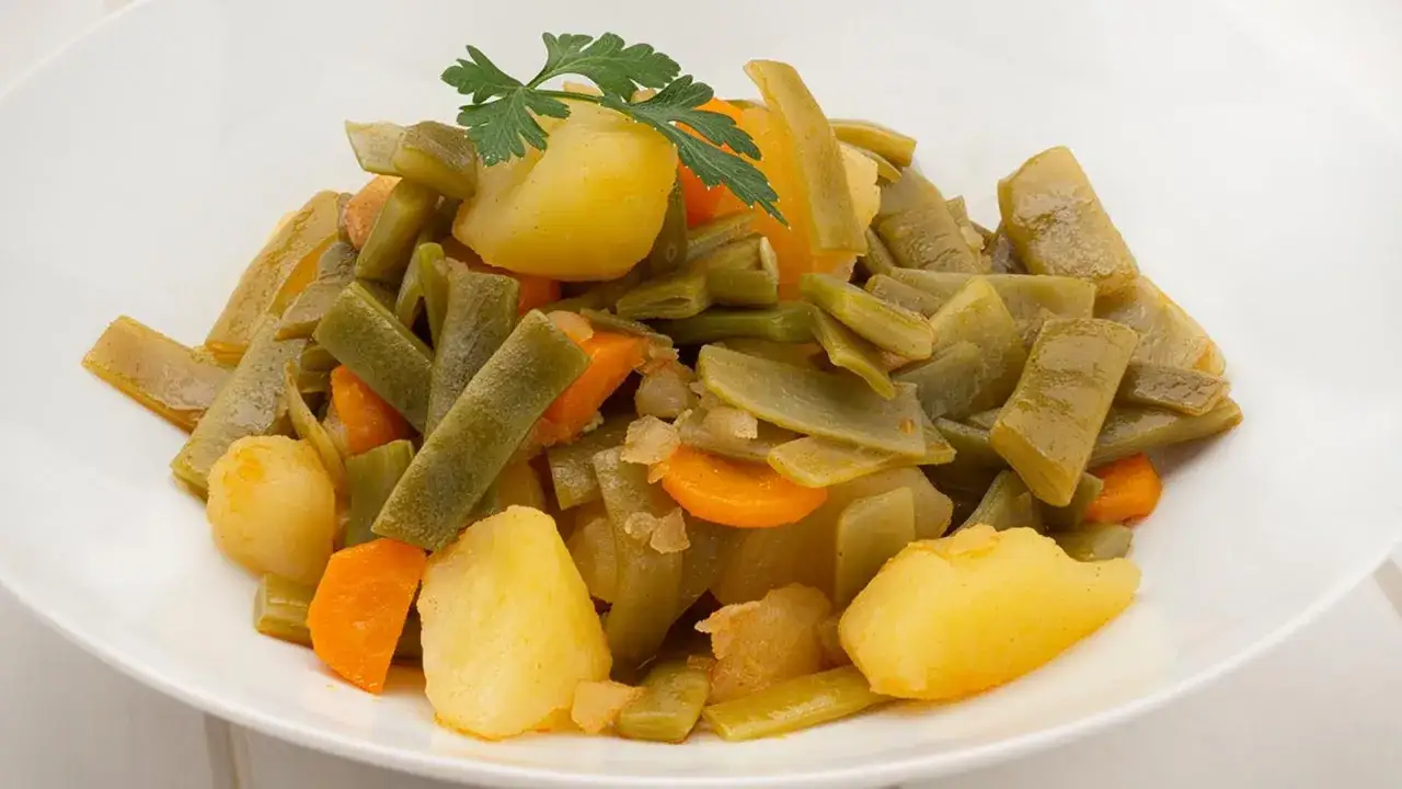 Un plato blanco lleno de judías verdes tiernas, trozos de patata dorada y rodajas de zanahoria, todo ello aderezado con una ramita de perejil fresco. Un guiso casero de judías con patatas.