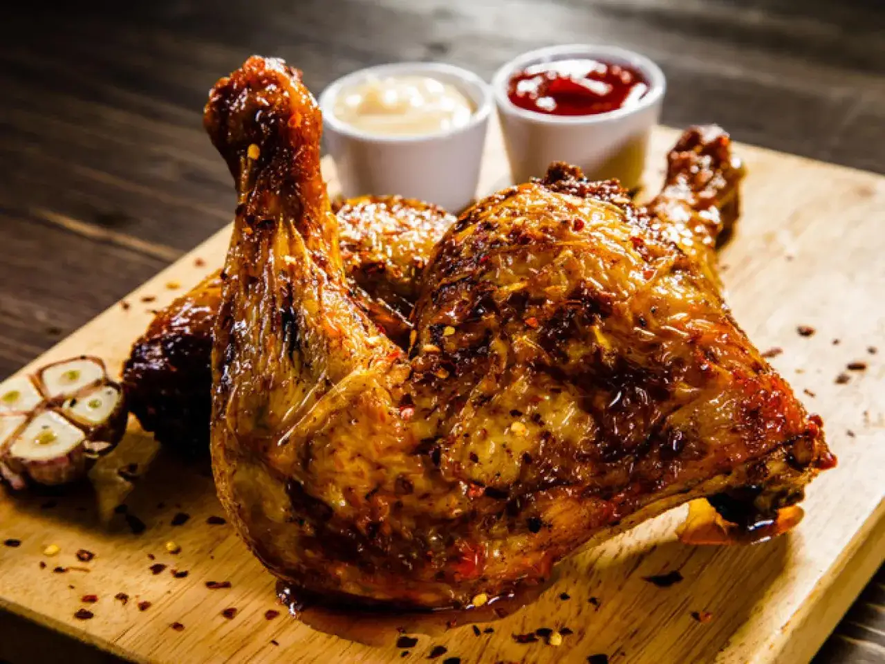 Contramuslos de pollo al horno crujientes, dorados y jugosos, servidos con salsas y ajo asado en tabla de madera.
