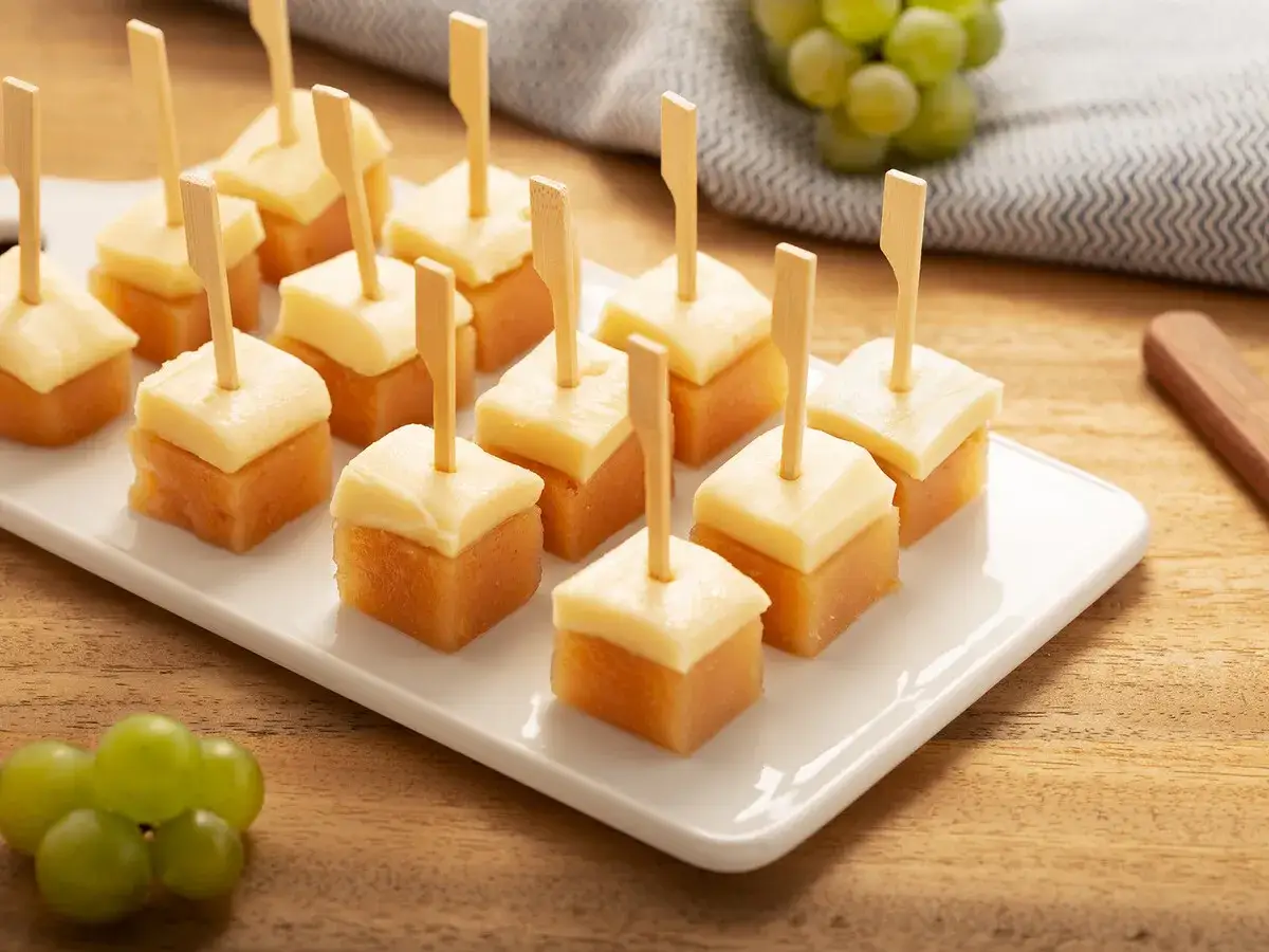 Bocaditos de queso y dulce de membrillo en palillos, servidos en una bandeja blanca.
