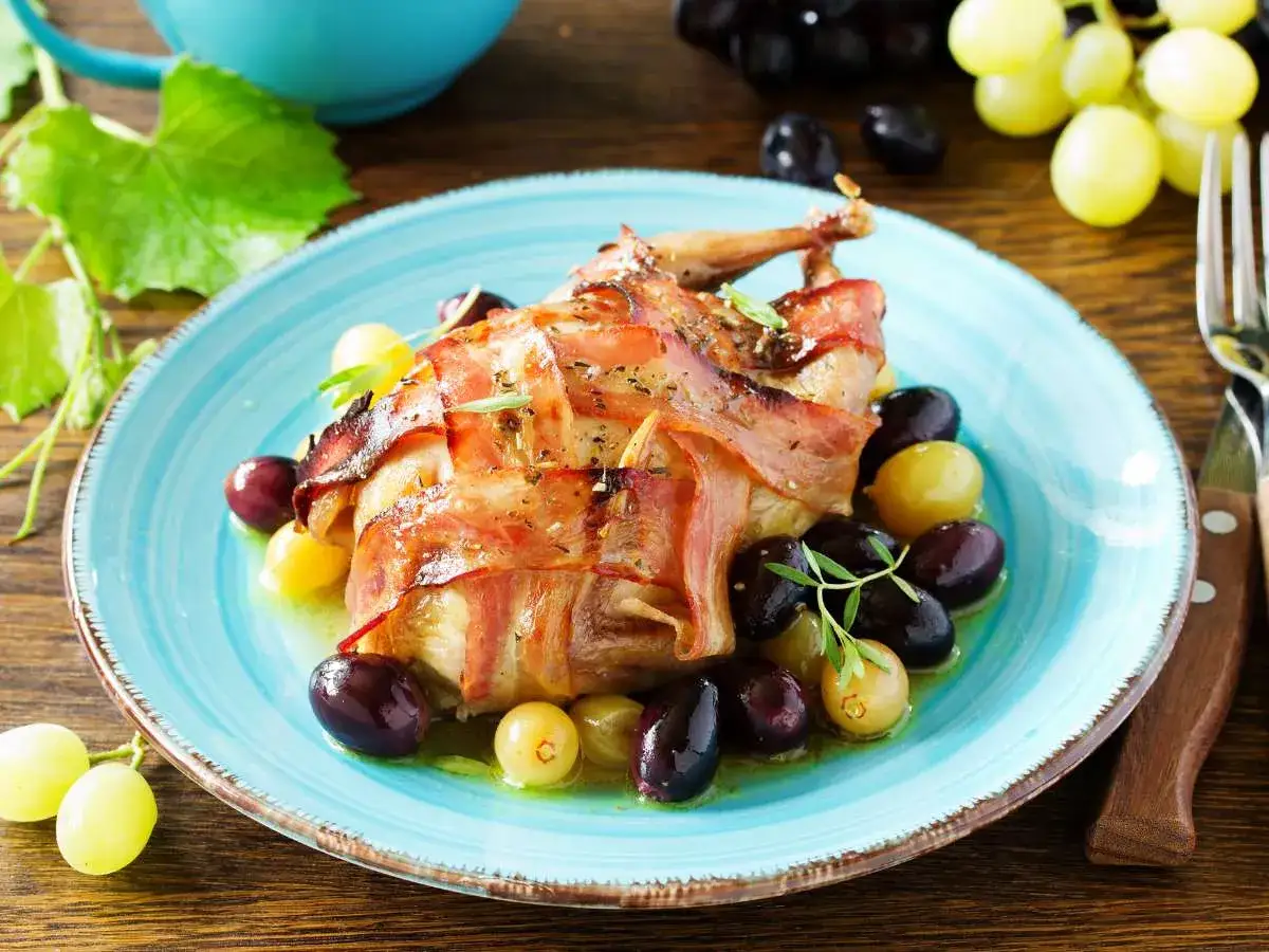 Deliciosas codornices al horno envueltas en tocino, servidas con uvas verdes y moradas en un plato azul.