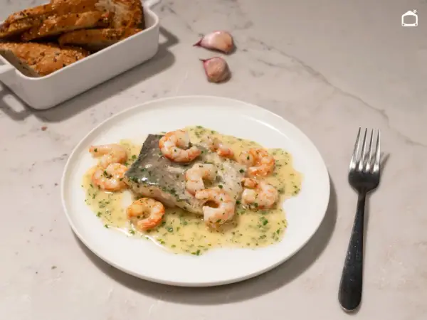 Deliciosa merluza con gambas en salsa cremosa, acompañada de pan rústico y ajos.