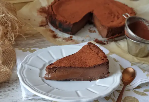 Una porción de tarta de chocolate, cubierta de cacao en polvo, reposa en un plato blanco. El resto de la tarta, también espolvoreada, se ve al fondo.