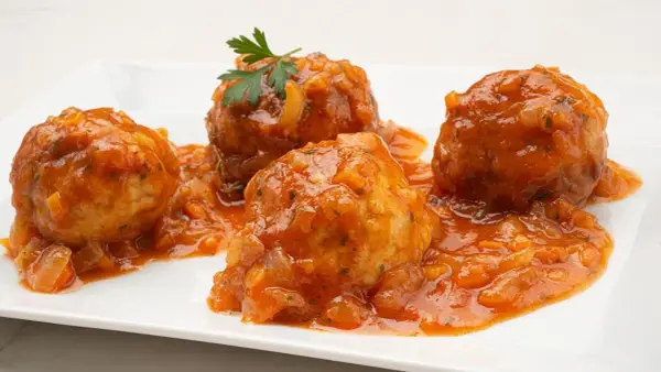Cuatro albóndigas de rape en una salsa de tomate casera, decoradas con perejil fresco.