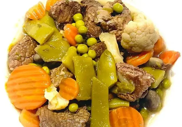 Ternera a la jardinera con trozos de carne tierna, zanahorias, guisantes, pimientos verdes y coliflor en un plato blanco.