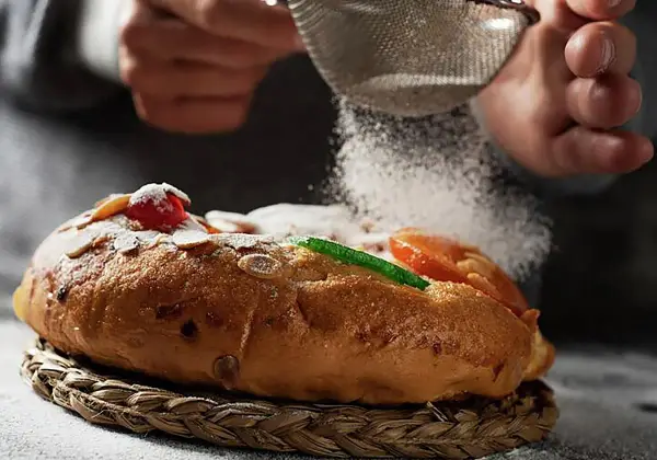 Manos espolvorean azúcar glas sobre un roscón de reyes decorado con frutas escarchadas y almendras. ¡Una delicia para tu roscón de reyes receta!